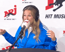 a woman wearing headphones and a blue jacket is speaking into a microphone in front of a hit music only sign