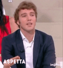 a man in a suit and white shirt is sitting on a couch with the word aspettta written on the bottom