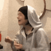 a woman wearing a hooded sweatshirt and a beanie is laughing in front of a mirror .