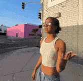 a woman wearing sunglasses and a white tank top stands on a sidewalk in front of a pink building