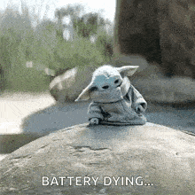 a baby yoda is sitting on top of a rock with the words `` battery dying '' written on it .
