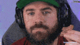 a man with a beard wearing headphones and a green hat is eating a snack