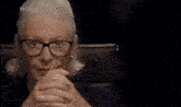 an older woman wearing glasses and earrings sits with her hands folded in front of her face