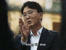 a man wearing glasses and a suit is clapping his hands with the words không tin được written below him .