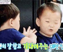 a baby in a blue shirt looks at himself in the mirror