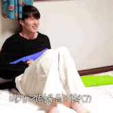 a man in a black sweater sits on a bed with korean writing on the bottom right corner