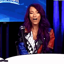 a woman with purple hair and a leather jacket is sitting at a table with her mouth open .