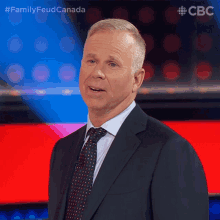 a man in a suit and tie stands in front of a red and blue background with the words family feud canada on the bottom