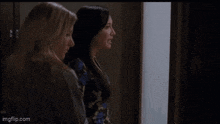 two women are standing next to each other in a room and looking out a window .