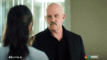 a man with a mustache is talking to a woman and the nbc logo can be seen behind him