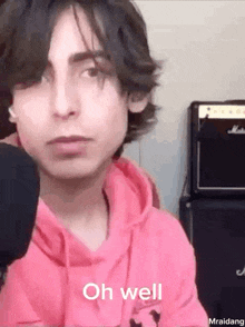 a young man in a pink hoodie is standing in front of a microphone and saying `` oh well '' .