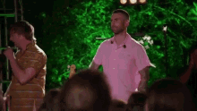 a man in a pink shirt is singing into a microphone while a man in a yellow shirt is playing drums .