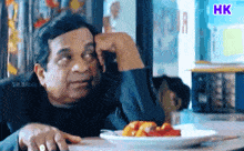 a man sits at a table with a plate of food in front of him with the letters hk on the bottom right