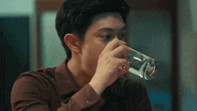 a man in a brown shirt is drinking water from a clear glass