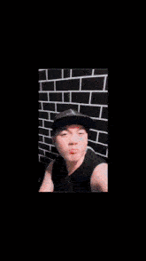a man standing in front of a brick wall wearing a black hat