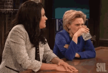 two women sit at a bar with snl written on the bottom right