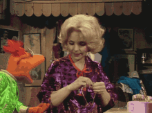 a woman in a purple top is knitting while a muppet in a green outfit looks on
