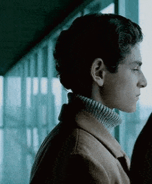 a young man wearing a turtleneck sweater and a brown jacket is looking out a window .