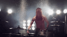 a shirtless man plays drums in front of a spotlight