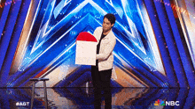 a man in a white jacket is holding a red white and blue object on a stage with a nbc logo in the background
