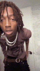 a young man with dreadlocks is standing in front of a door wearing a purple shirt and black pants .