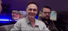 a man in a denim jacket is smiling while sitting next to another man in glasses .