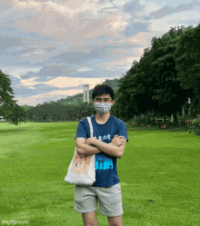 a man wearing a mask stands in a field with his arms crossed and a tote bag