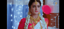 a woman in a red and white saree and gold jewelry is standing in front of a window .