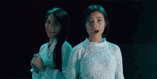 two women are standing next to each other in a dark room . one of the women is wearing a white lace shirt .