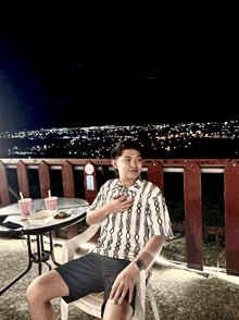 a man in a striped shirt sits in a white chair with a city in the background