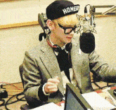 a man wearing a hat that says homer sits at a desk in front of a microphone