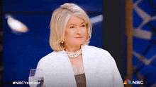 a woman is sitting at a table with a glass of water and a nbc logo behind her