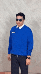a man wearing sunglasses and a blue sweatshirt is standing in front of a wall .