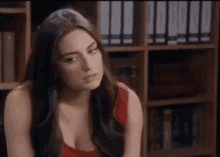 a woman in a red tank top is sitting in front of a bookshelf and looking at the camera .