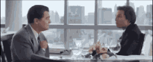 two men are sitting at a table in front of a window talking