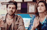 a man and a woman are sitting next to each other in a room and the woman is holding her hands to her chest .