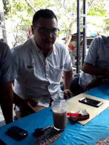 a man wearing glasses sits at a table with a drink and a pack of cigarettes