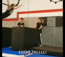 a woman is doing a trick on a trampoline in a gym while a man watches .