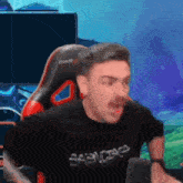 a man with a mustache is sitting in a red and black gaming chair and making a funny face .
