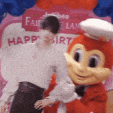 a woman stands next to a jollibee mascot in front of a sign that says happy birthday