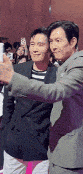 two men in suits are taking a selfie together in front of a crowd of people .