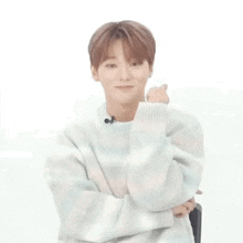 a young man in a sweater is sitting in a chair with his arms crossed and making a heart shape with his hand .