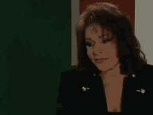 a woman wearing a black jacket and a plunging neckline is sitting in front of a green wall .