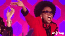 a woman in a red jacket and sunglasses is dancing with her arms in the air and shouting .