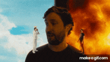 a man is standing in front of a fire while another man stands in front of a cloudy sky