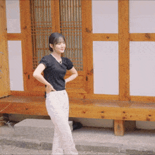 a woman wearing a black top and white pants stands in front of a wooden building