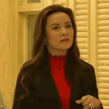 a woman wearing a red sweater and a black jacket is standing in front of a white wall .