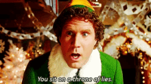a man in a green elf costume is sitting on a throne of lies with a christmas tree in the background .