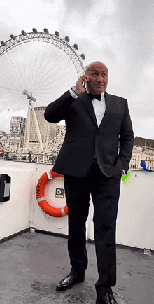 a man in a tuxedo talks on a cell phone in front of a large ferris wheel