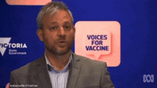 a man in a suit stands in front of a sign that says " voices for vaccine "